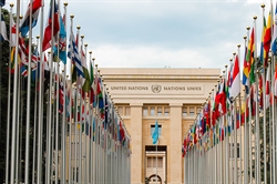 [ai] The building of the United Nations in Geneva, viewed from a pathway lined with flags of various countries, under a cloudy sky.